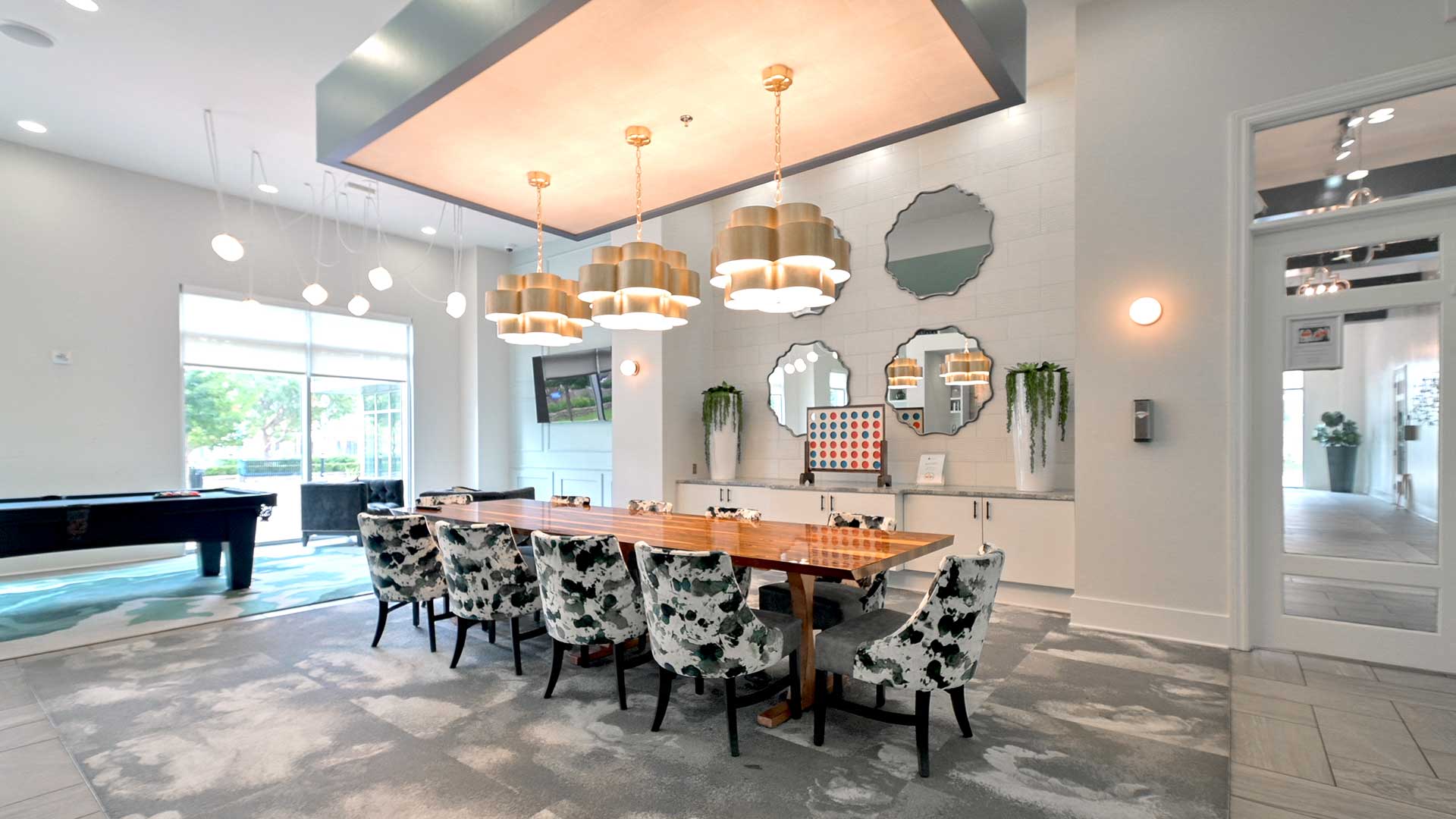 A large dining table with pattered chairs sits below three, large chandeliers. Behind is a countertop with a game and potted plants on it. To the left is a pool table and windows looking out to the po