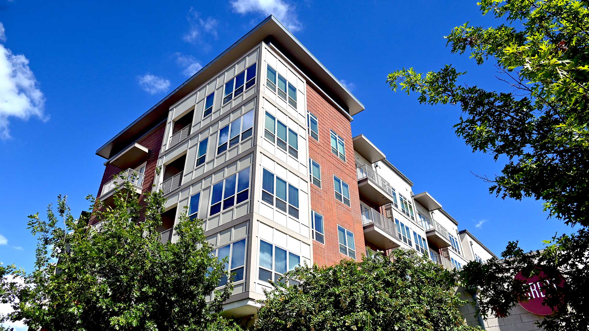 Looking up at the exterior corner of the apartment building. The façade is part beige, part red brick. There are trees at the base and the sky is blue. The Crest at Las Colinas office sign can be seen