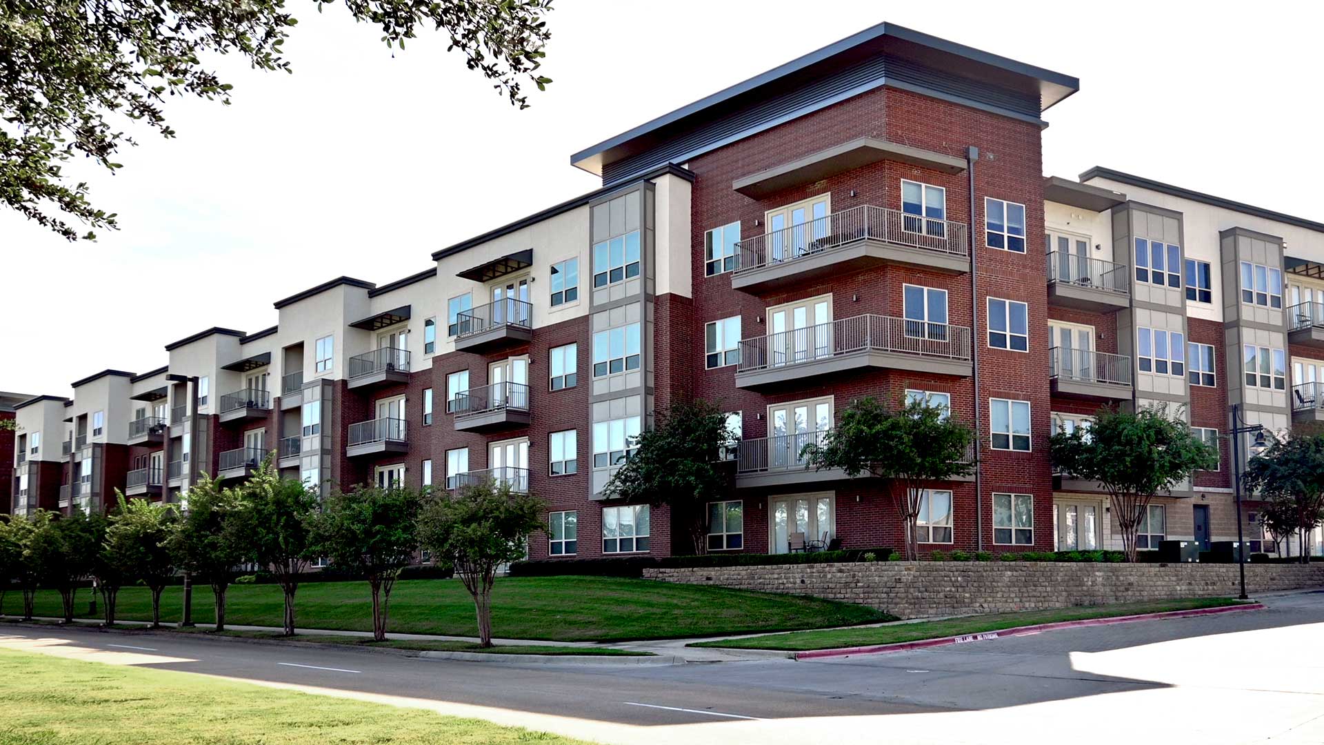 Looking at the corner of the apartment building from across the street. The building is primarily red brick with beige sections. The corner has balconies that wrap around the corner. The street is lin