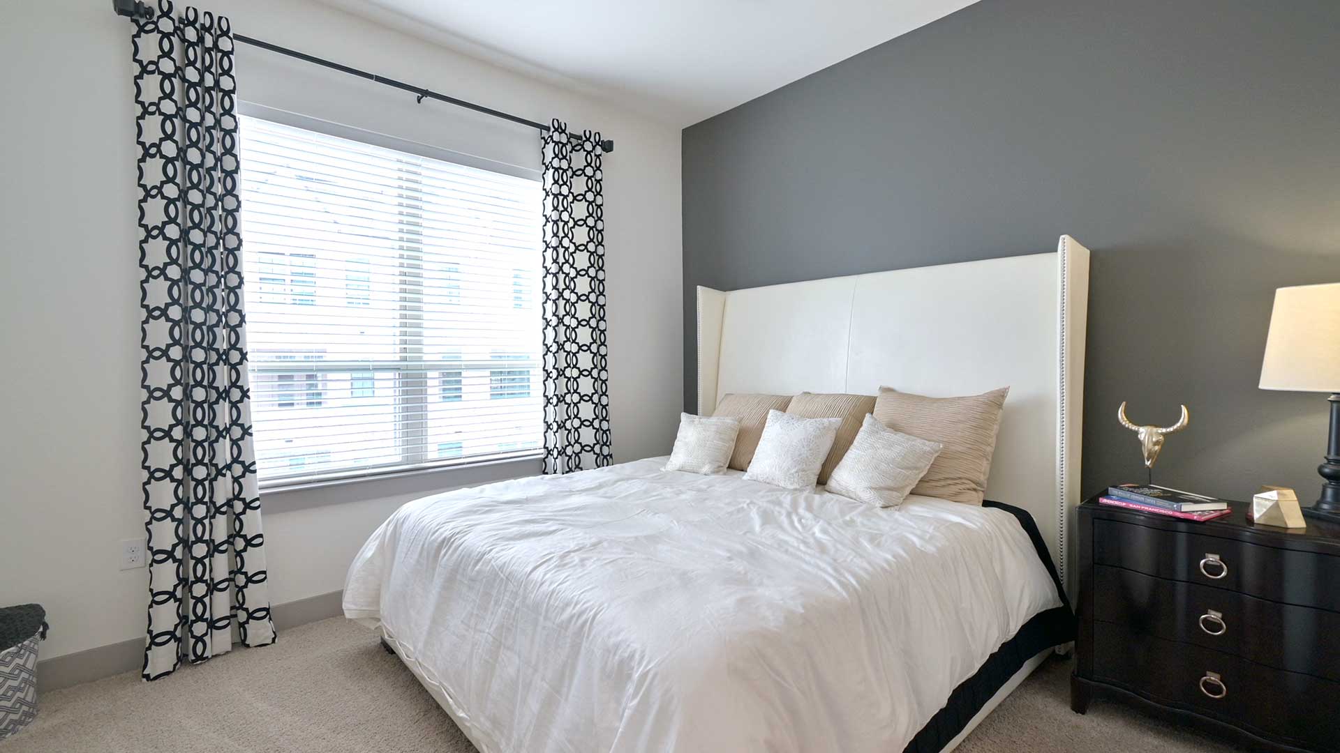 A large bed in a carpeted bedroom. Curtains are open hanging on the window to the left and a dresser is seen next to the bed on the right.