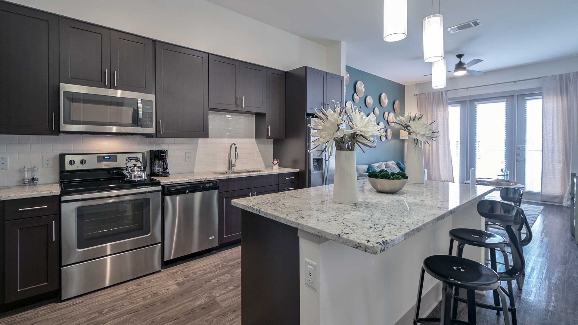 Looking along the kitchen island in a residence at Crest at Las Colinas Station. The kitchen runs along the left wall with ebony cabinets and stainless steel appliances. The island has two large flowe