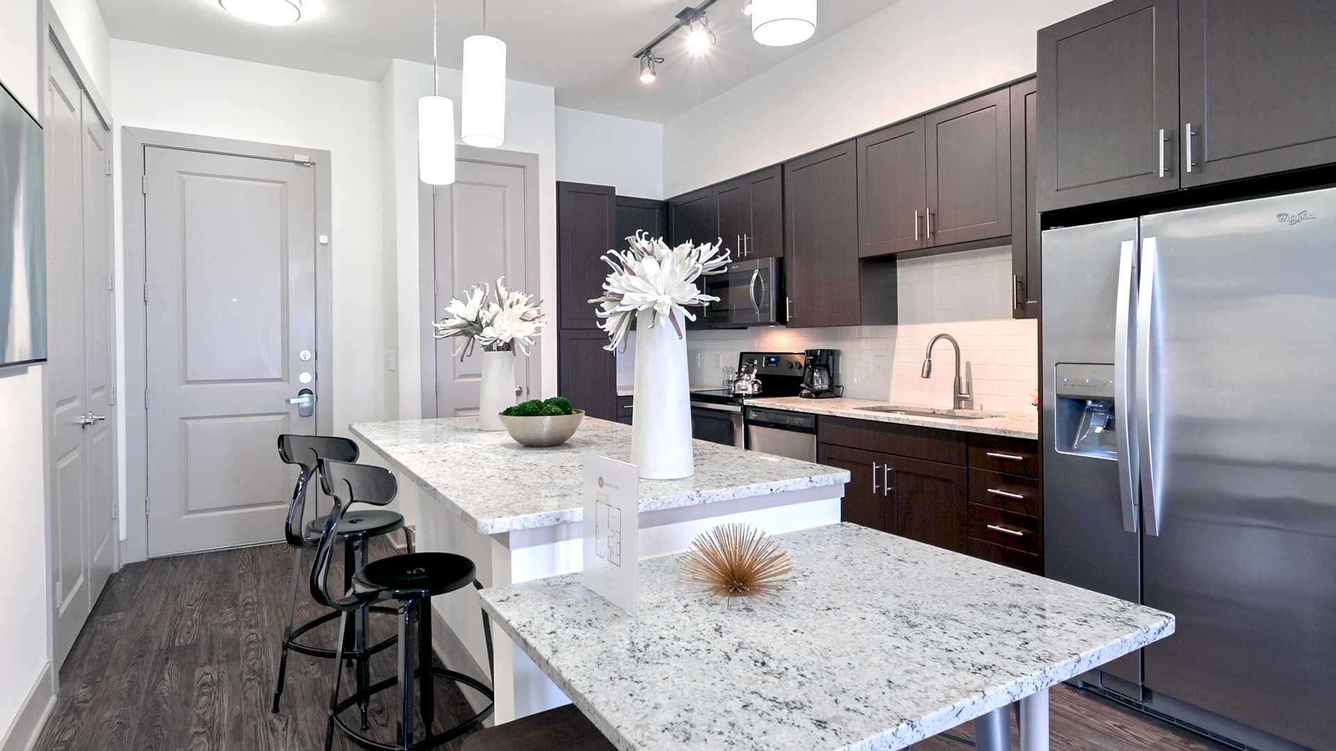 Looking across the kitchen island in a residence at Crest at Las Colinas Station. There are two stools in front of the island to the left with the front door behind.