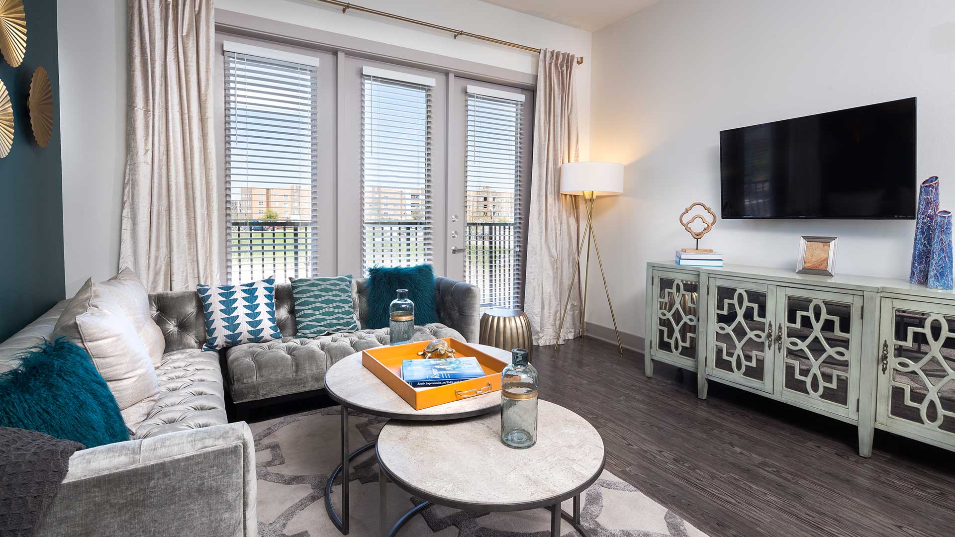 A living room in a residence at Crest at Las Colinas Station. A sectional couch is on the left with a set of round coffee tables before it. On the right is a dresser with various art pieces on it and