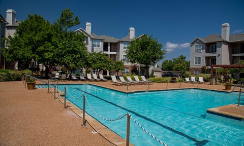 A pool surrounded by chairs and trees with apartment buildings in the background.