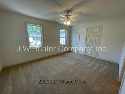 A room with a carpet floor, a ceiling fan, and a window showing a view of trees and a building.