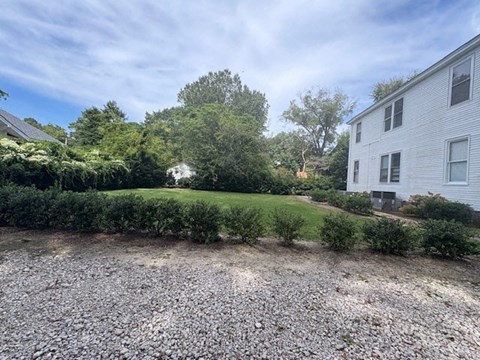 A gravel area in front of a white house with a green hedge and trees in the background.