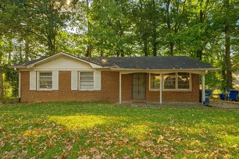 A small house with a brown brick exterior and a grey roof surrounded by trees.