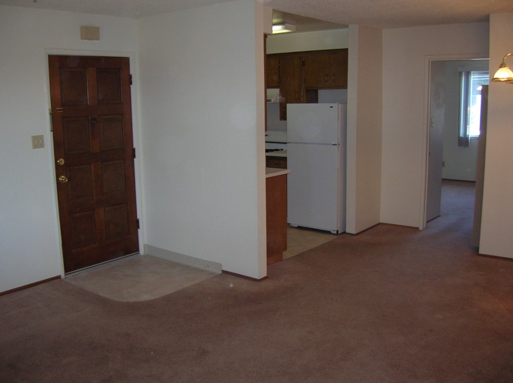 an empty living room with a door to a kitchen and a refrigerator
