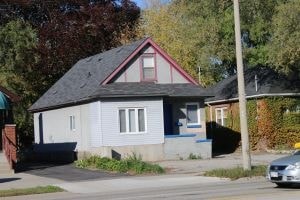 A small house with a red roof and a grey body is situated on a street.