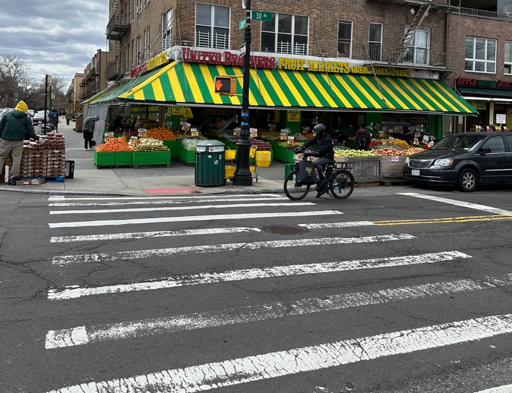 A man on a motorcycle is crossing the street in front of a produce stand.