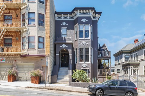 A grey car is parked in front of a two-story building with a balcony.
