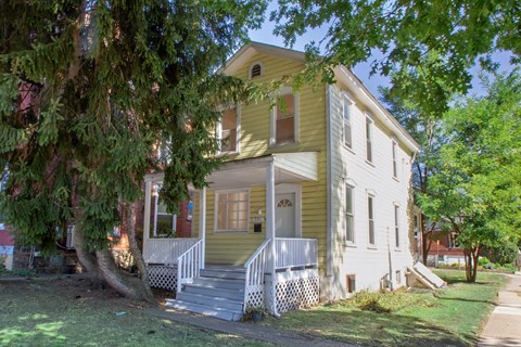 A yellow house with a white porch and a tree in front.