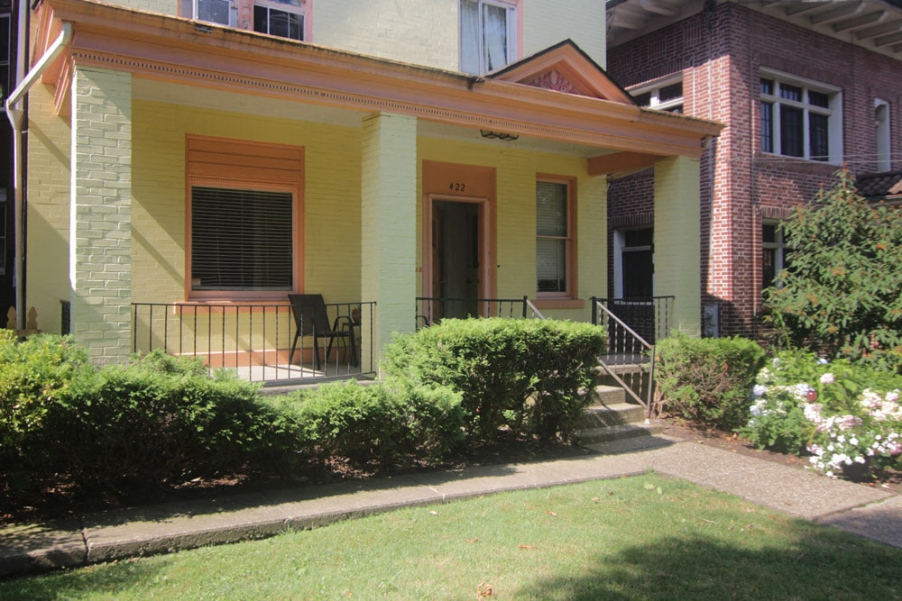 a front porch of a yellow house with a lawn and bushes