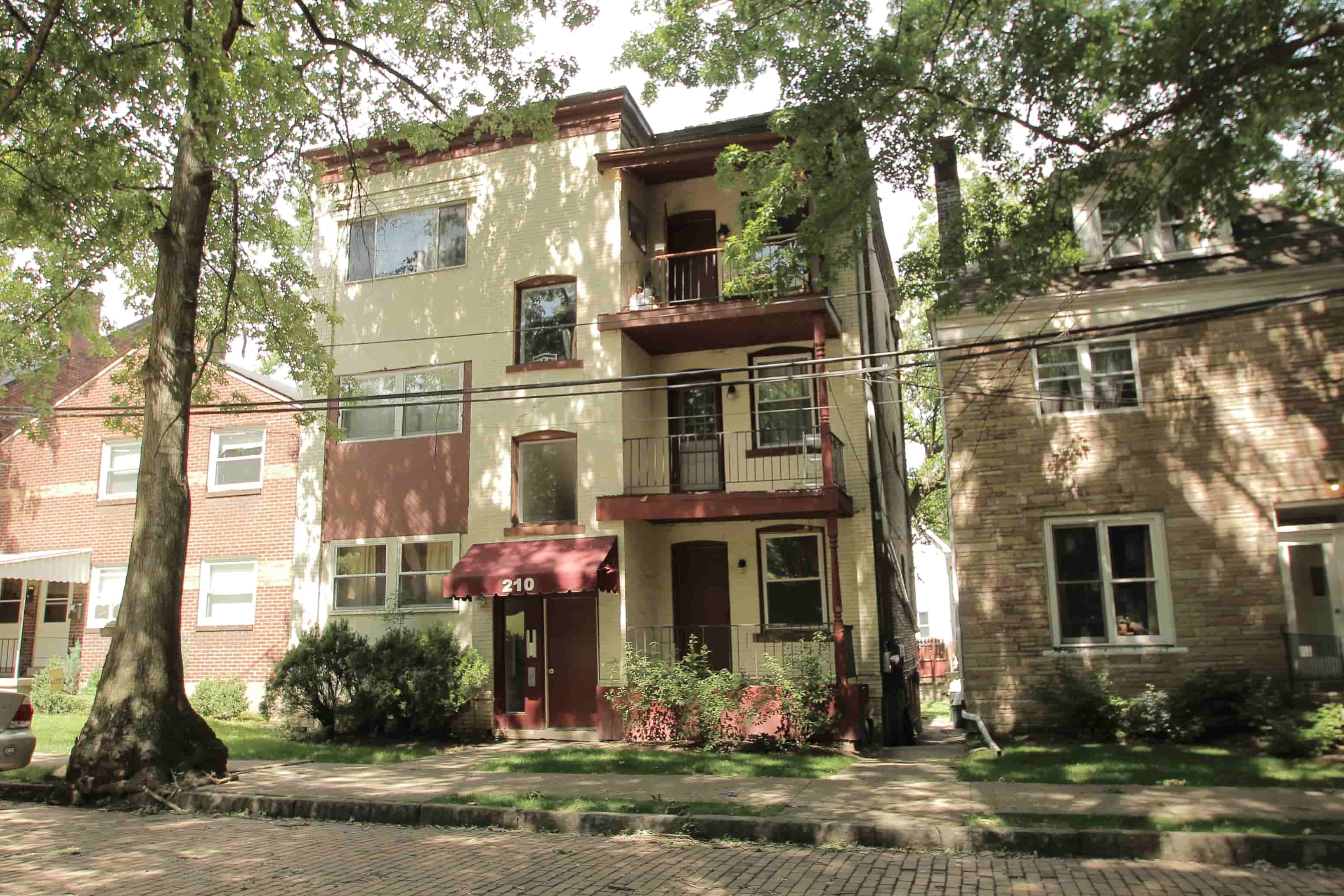 A building with a balcony and a red awning is surrounded by trees and bushes.