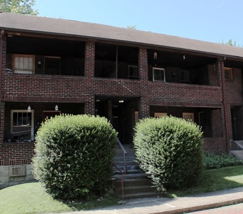 A red brick building with a porch and two bushes in front.