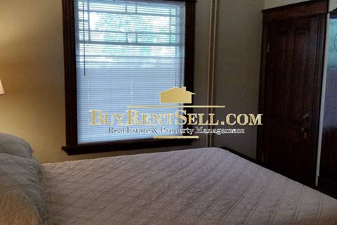 A bedroom with a bed in the foreground and a window with a sign that says BuyKentSell.com Real Estate & Property Management.