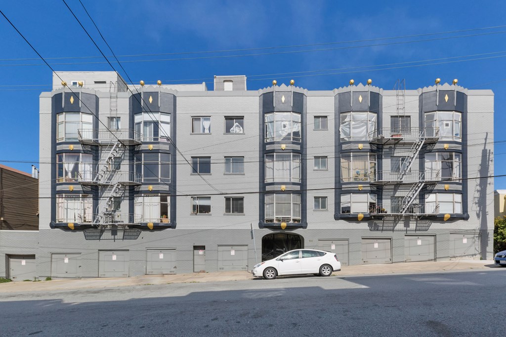 A white car is parked in front of a grey building with a fire escape.