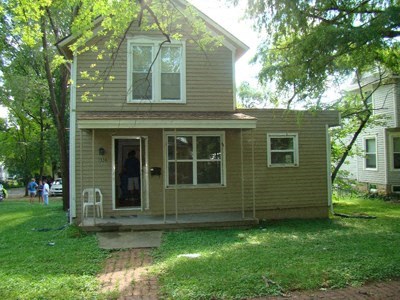 A house with a brown siding and a white door is surrounded by trees.