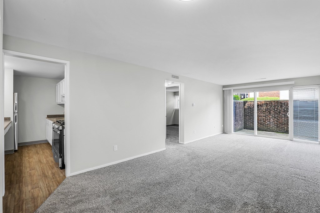 A spacious living room with a grey carpet and a view of a fenced area outside the window.