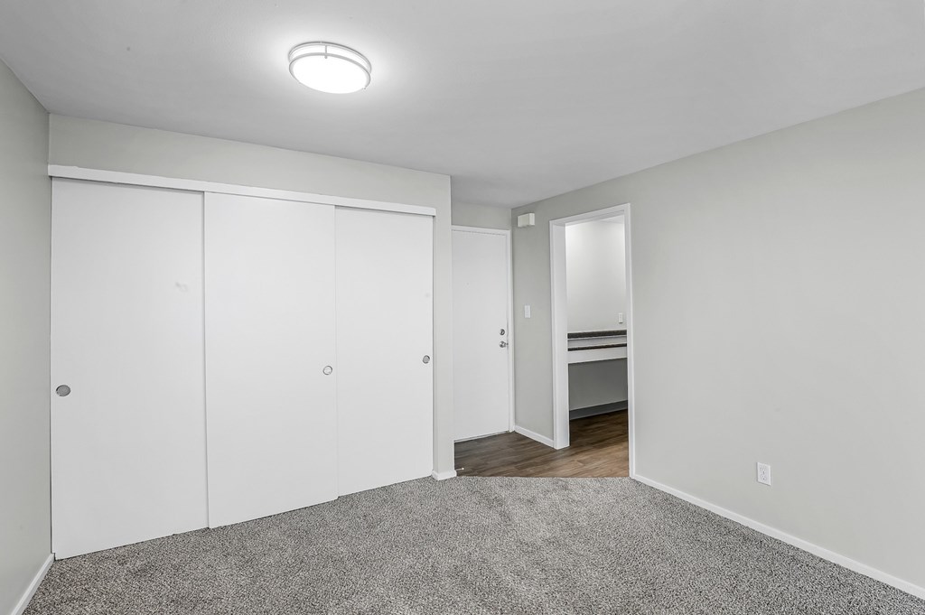 A bedroom with a white door, a grey carpet and a white wall.