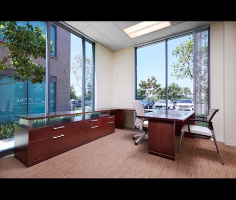 A room with a desk and chairs with a view of trees and a building outside the window.