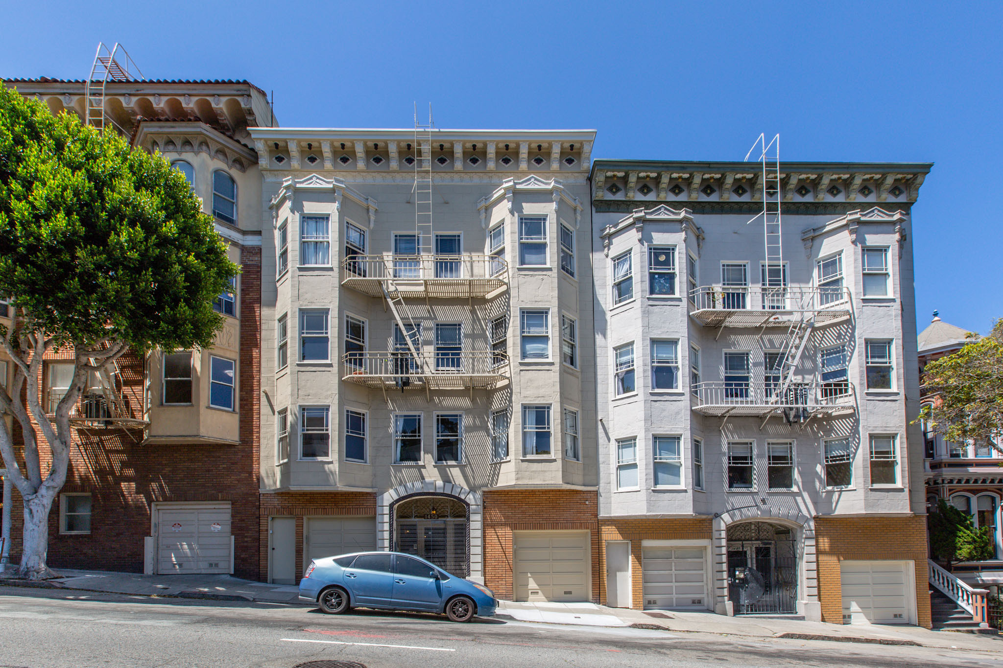 A blue car is parked in front of a building with a tree on the left.