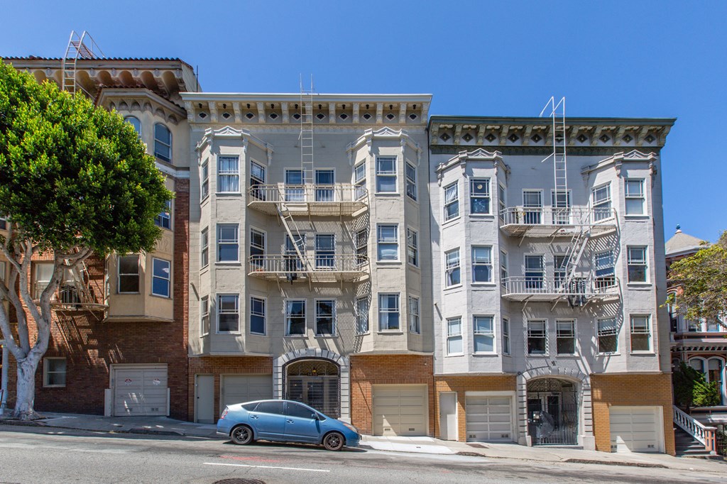 A blue car is parked in front of a building with a balcony.