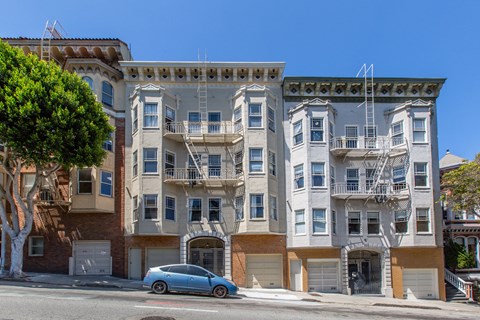 A blue car is parked in front of a building with a balcony.