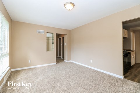 A spacious room with a carpeted floor and a kitchen area visible in the background.