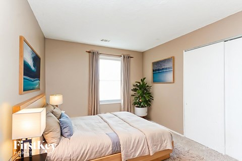A bedroom with a large bed, a nightstand with a lamp, and a plant on the windowsill.