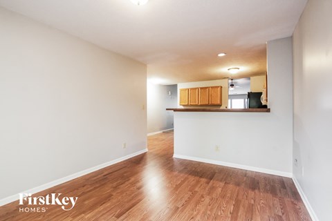 A room with wooden floors and white walls, with a kitchen area visible in the background.