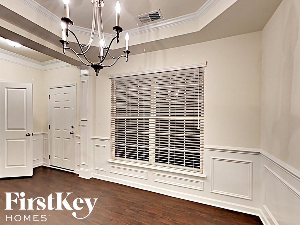 A chandelier hangs over a window with white blinds in a room with wooden floors and white walls.
