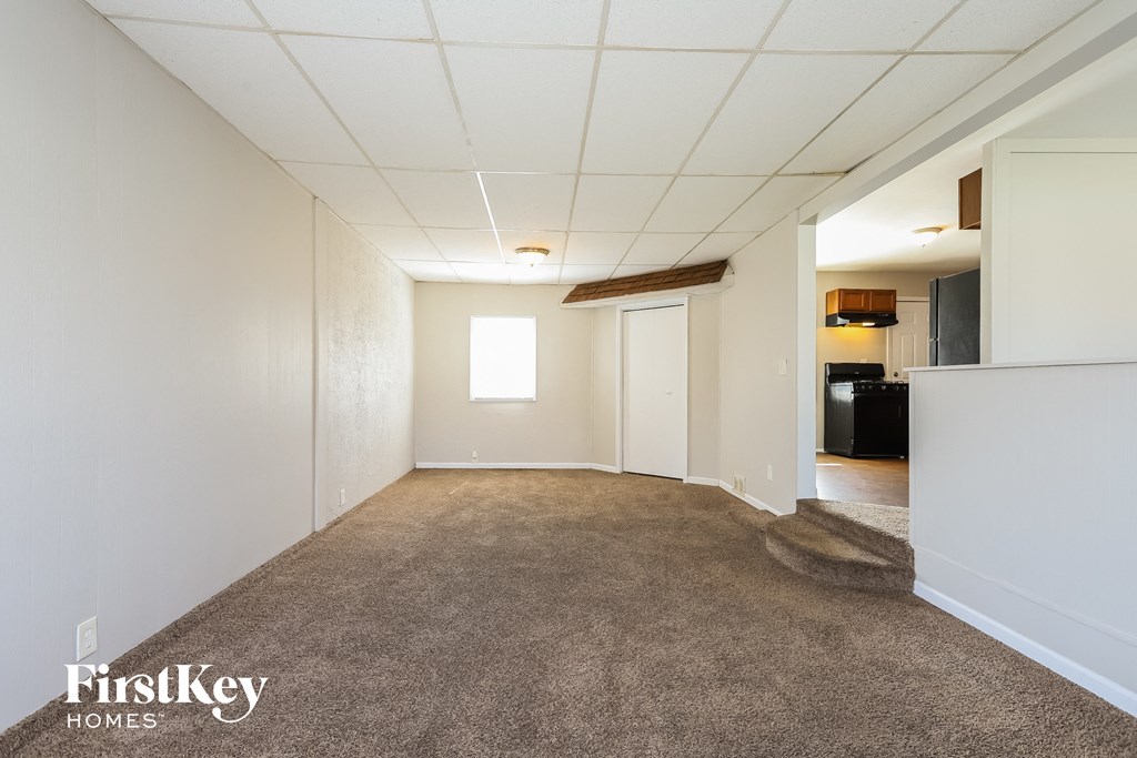 A room with a carpeted floor and white walls, with a door on the left and a kitchen area on the right.