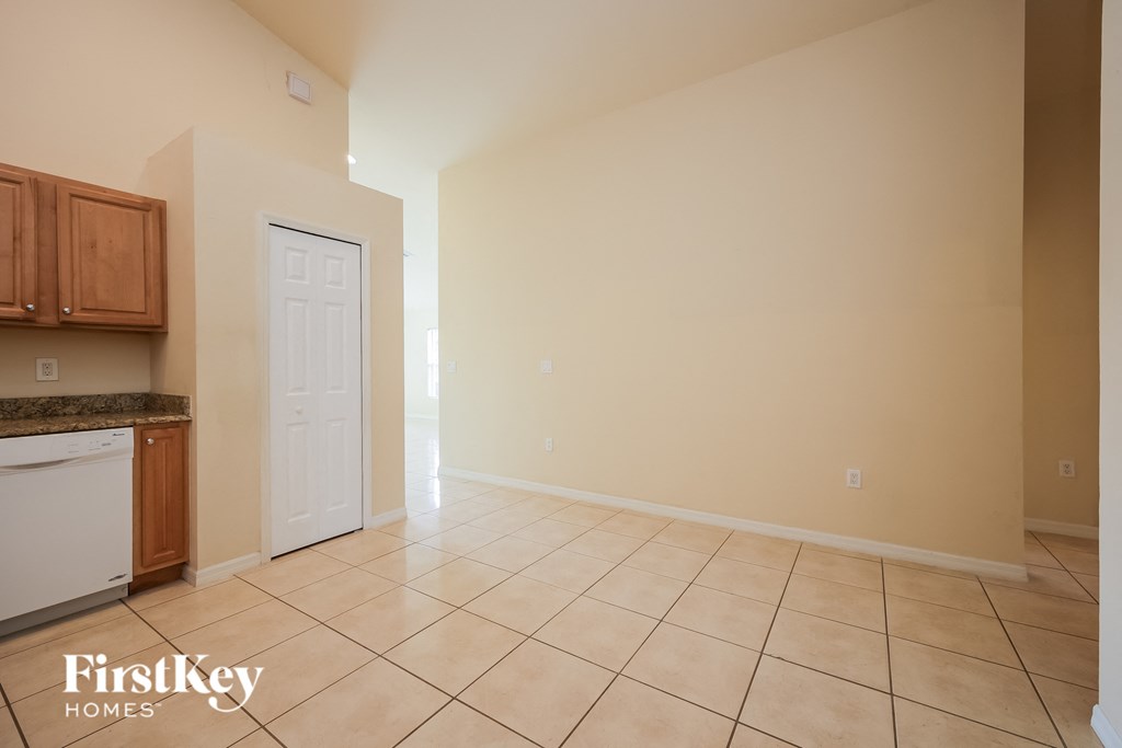 A spacious room with beige walls and tiled flooring, featuring a kitchen area with wooden cabinets and a white dishwasher.