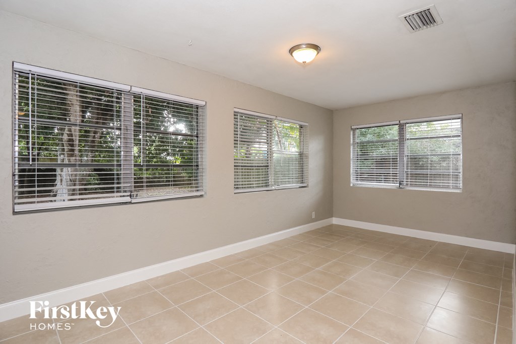 A room with beige walls and tiled flooring, with three windows covered in blinds.
