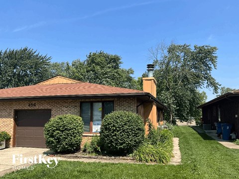 A house with a brown roof and a chimney is surrounded by green bushes and trees.