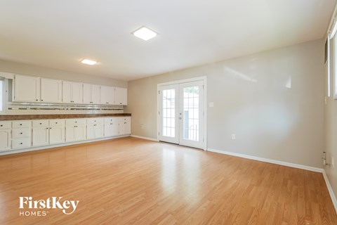 A spacious room with wooden floors and white walls, featuring a kitchen area with white cabinets and a window with blinds.