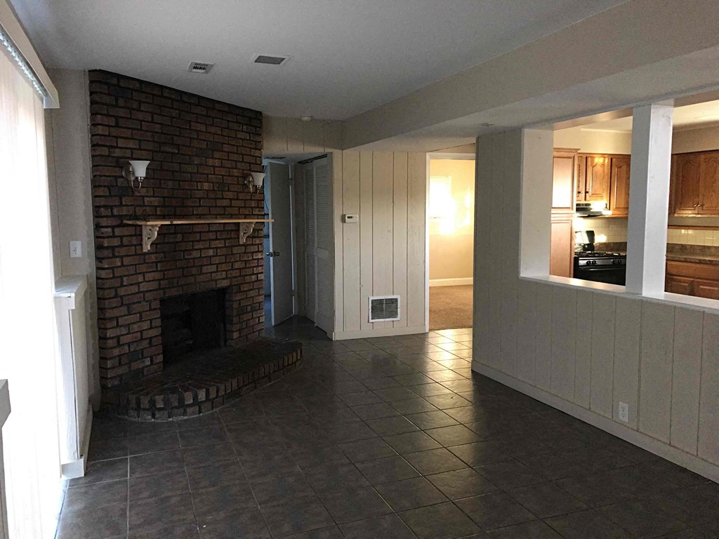 A living room with a brick fireplace and a kitchen in the background.