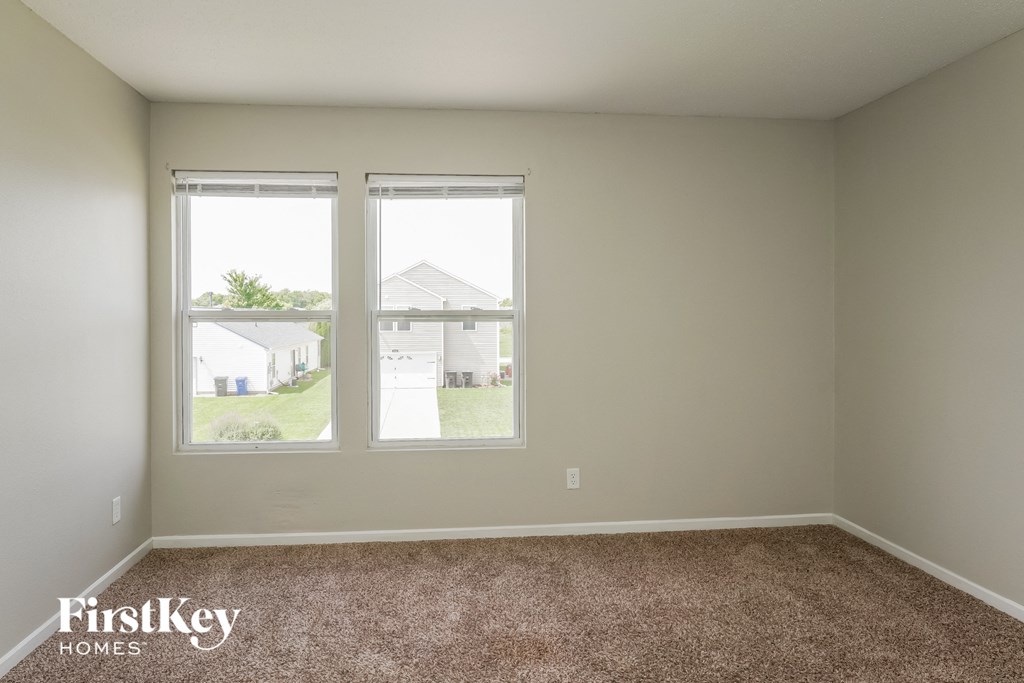 A room with a carpeted floor and two windows overlooking a residential area.