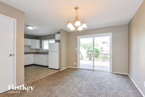A spacious living room with a kitchen in the background and a grey carpet on the floor.