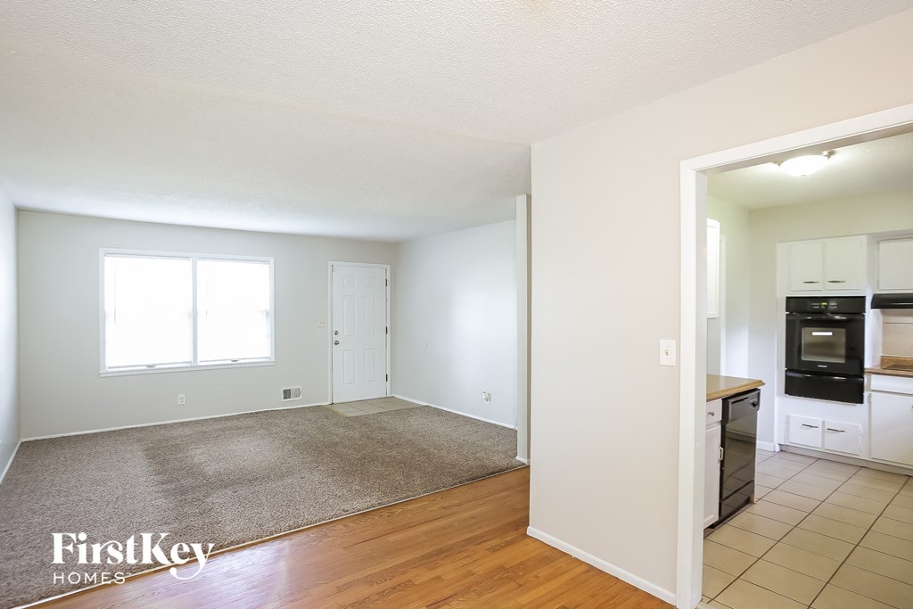 A room with a carpet and a kitchen in the background.