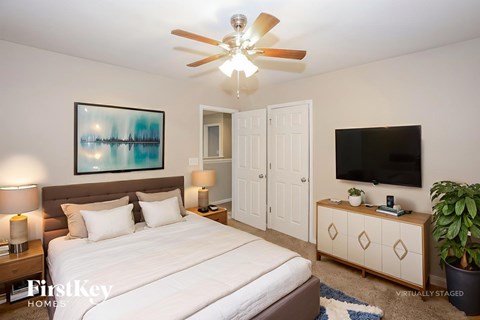 A bedroom with a bed, a ceiling fan, a nightstand with a lamp, a painting on the wall, a TV on a cabinet, and a potted plant.