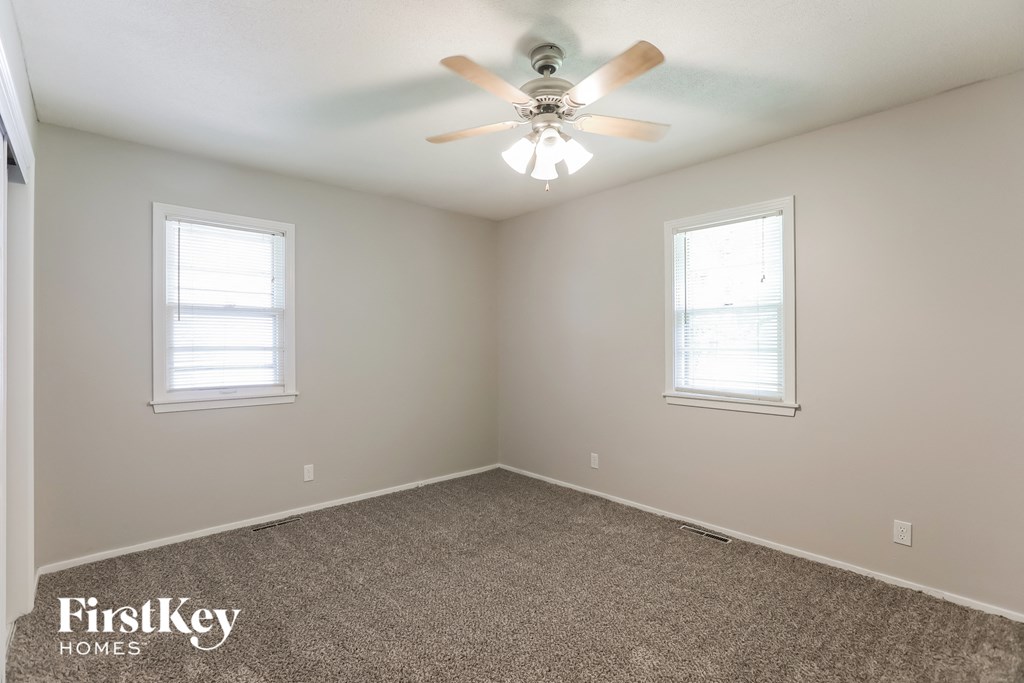 A room with a carpeted floor, two windows, and a ceiling fan.