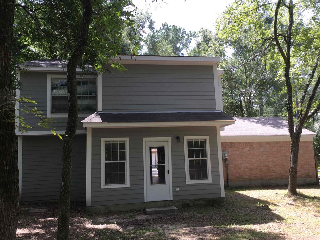 A small house with a grey siding and a white door is surrounded by trees.
