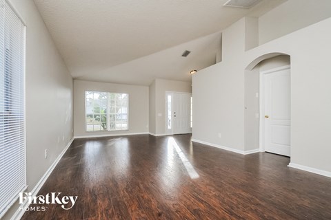 A spacious room with wooden floors and white walls, with a window on the left side.