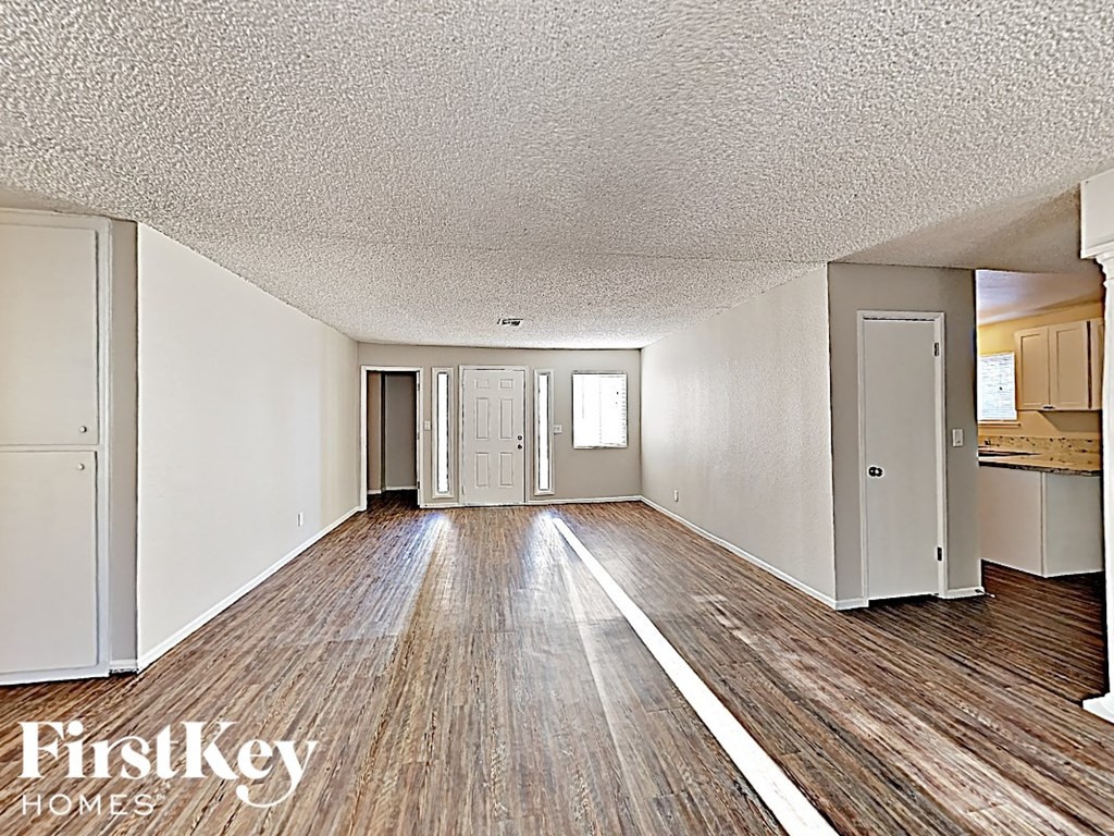 A room with wooden floors and white walls, with a kitchen area visible in the background.