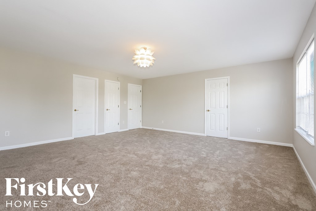 A spacious room with beige carpeting and white walls, with a light fixture on the ceiling and three white doors.