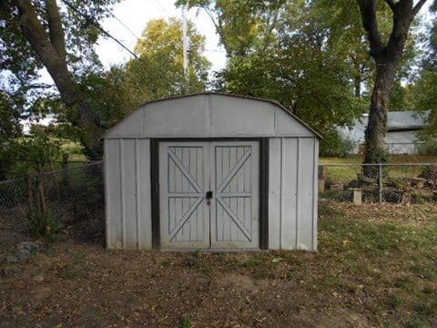 A white shed with a grey roof is surrounded by trees.