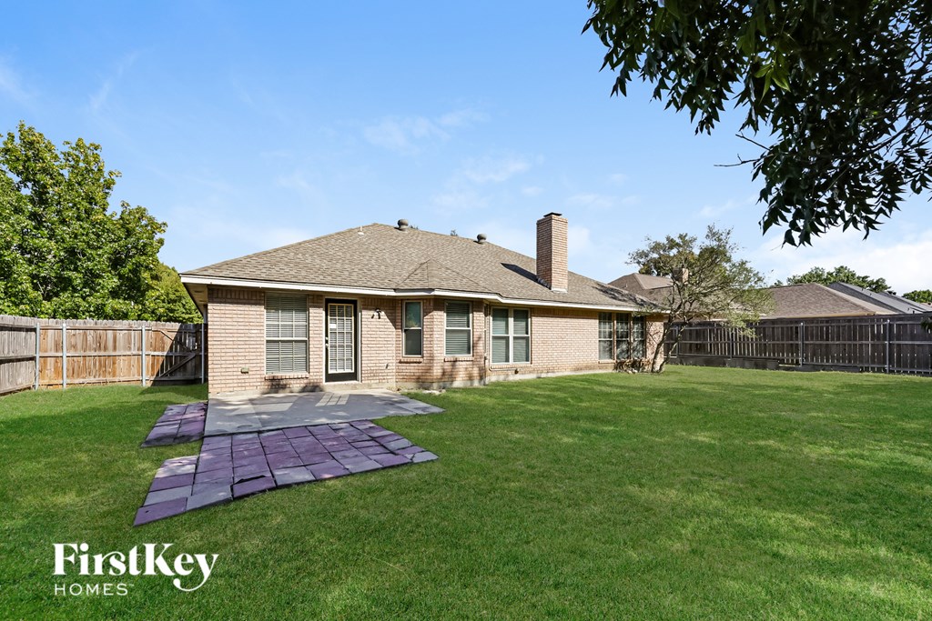 A house with a brown roof and a brick chimney is surrounded by a green lawn and a wooden fence.