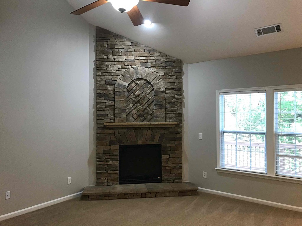A stone fireplace with a black firebox sits under a window in a room with a carpeted floor.